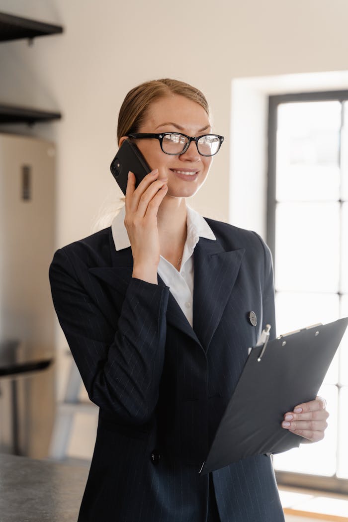 team-04 Smiling professional woman in business attire talking on phone holding clipboard in bright office.